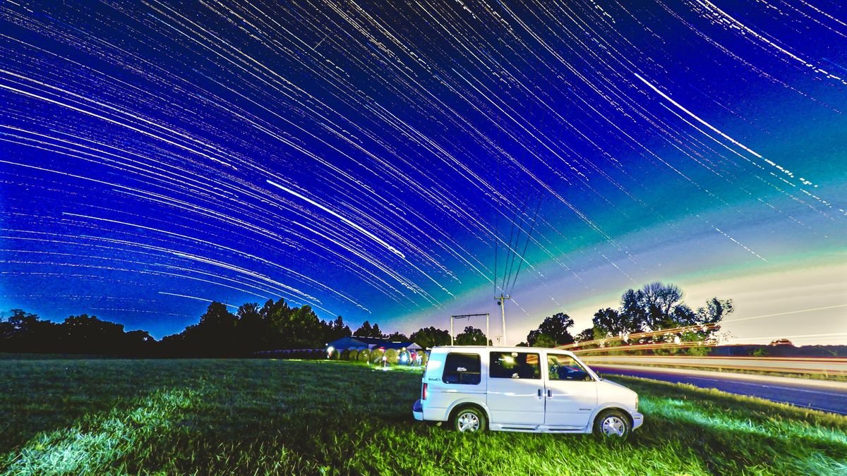 a white van is parked in a grassy field next to a road lined with the streaking lights of a passing car the sky is filled with the trails of stars