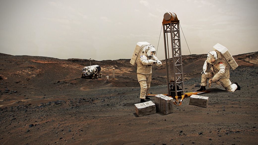 astronauts in space suits construct a short tower on the brownred surface of mars supply boxes lay about a rover is parked in the distance