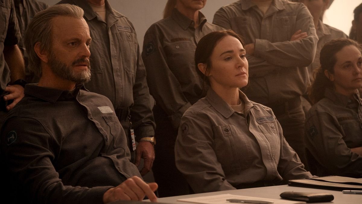 Sitting at a table are three people with very serious expressions In the background there are more people standing some with their arms crossed They are all wearing the same gray jumpsuit