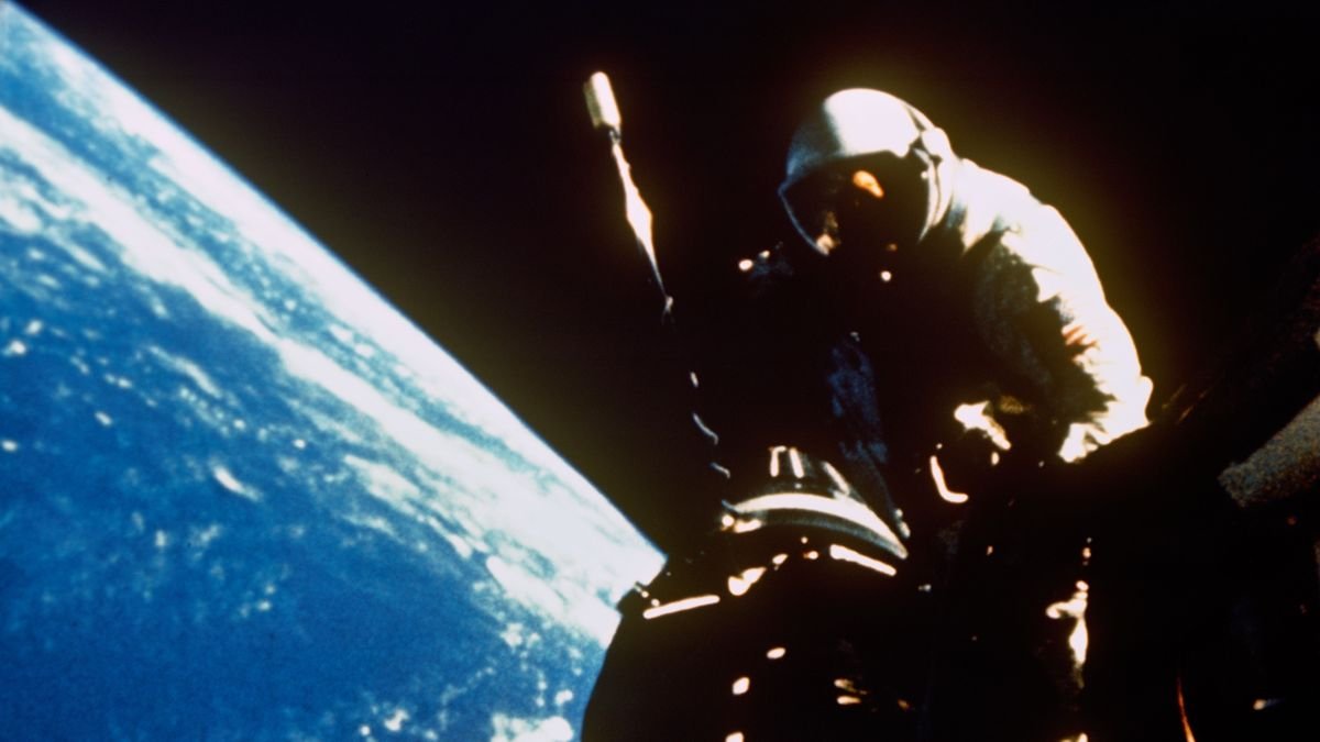 an astronaut floats near a shadowed spacecraft hatch against the black of space Part of Earth is seen below