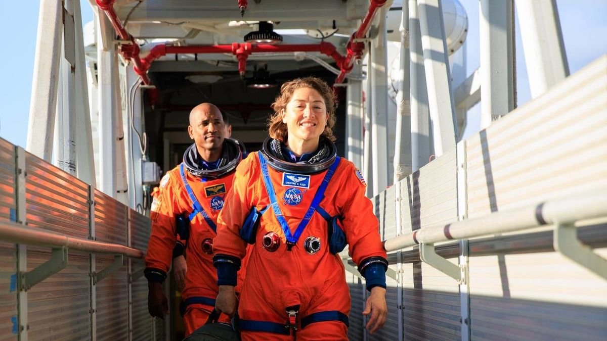 two humans in ornage space suits without helmets walk down a gantry way outside during a sunny day