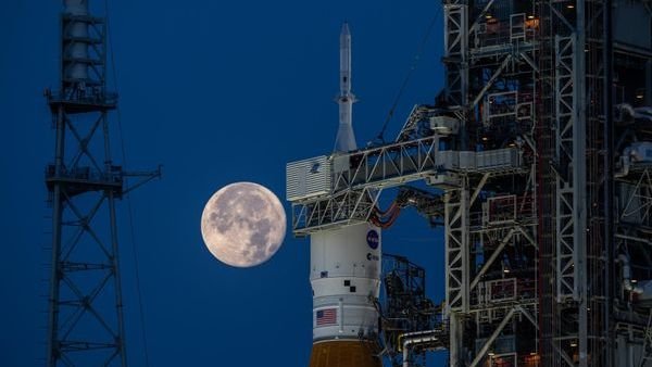the full moon hangs in frame with the orion inside its payload shell atop the SLS rocket