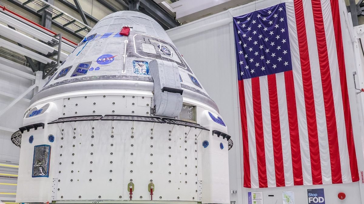 a silver and white space capsule sits inside a huge white walled room with an american flag in the background