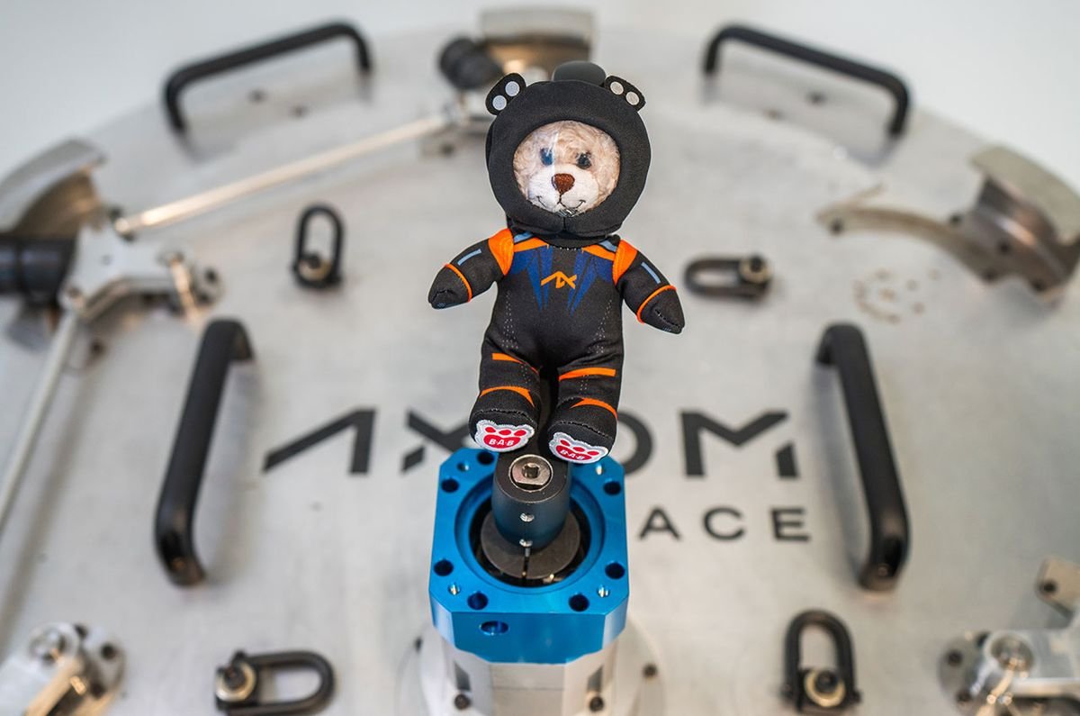 a small stuffed bear in a black spacesuit floats in front of white spacecraft hatch