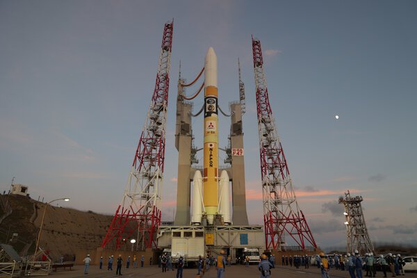 A rocket is rolled out to the launch pad at dusk with a crescent moon visible in the sky