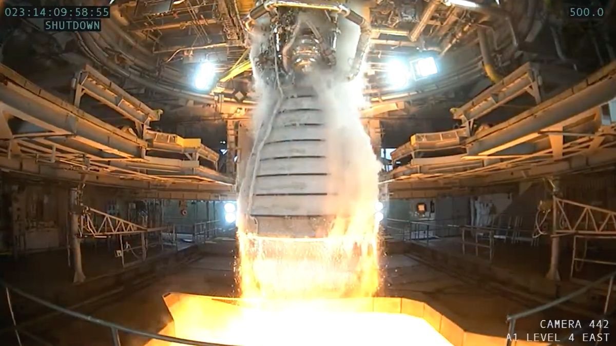 close up of a rocket engine with steam pouring out the sides and fire at the bottom surrounding is a large test stand