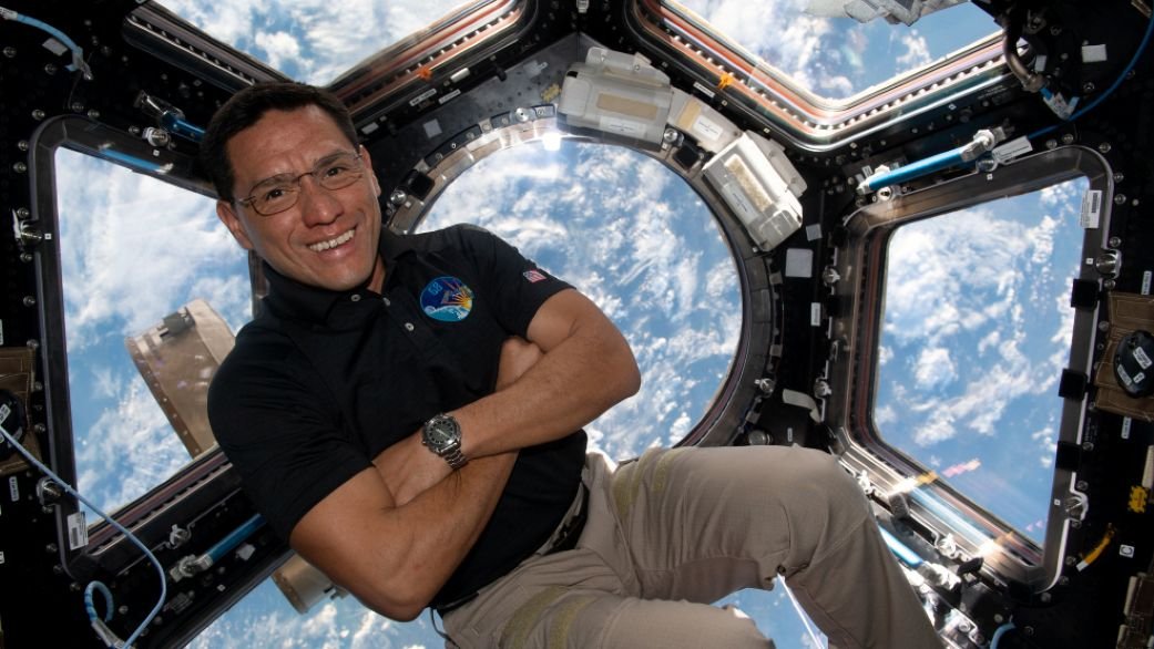A man in khakis crosses his arms as he floats in front of a hexagonal window that looks down at Earth from orbit