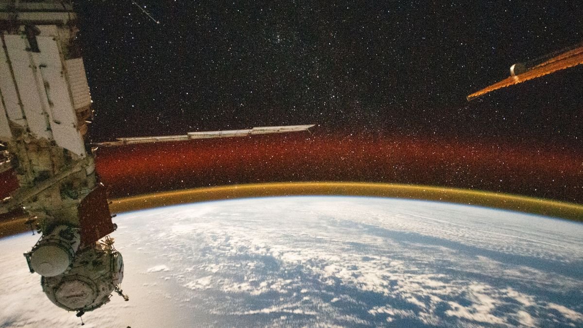 the round edge of earth is seen blue with a swath of clouds beneath a thin orange layer of atmosphere crowned in a red glow with the black starry sky of space above in the foreground on the left a russian space station capsule hangs downward On the right the golden orange reflective edge ot a solar panel slices the stars