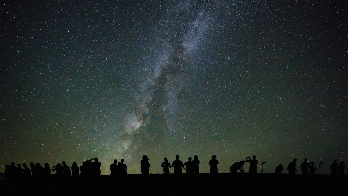 Silhouettes of people are seen standing on the ground looking up at a starry sky and the center of the Milky Way