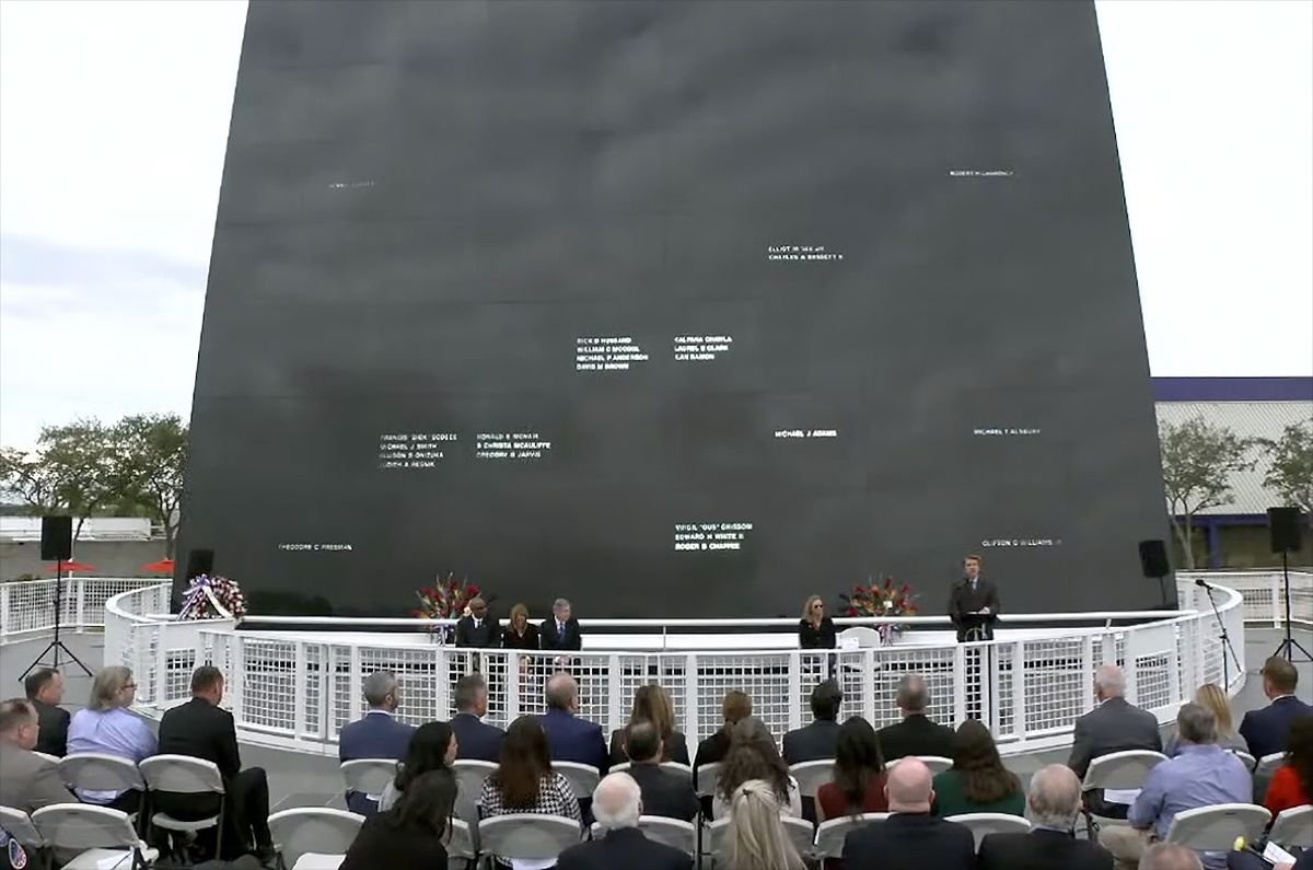 a handful of people in dark suits stand in front of a large black wall into which several dozen names have been carved