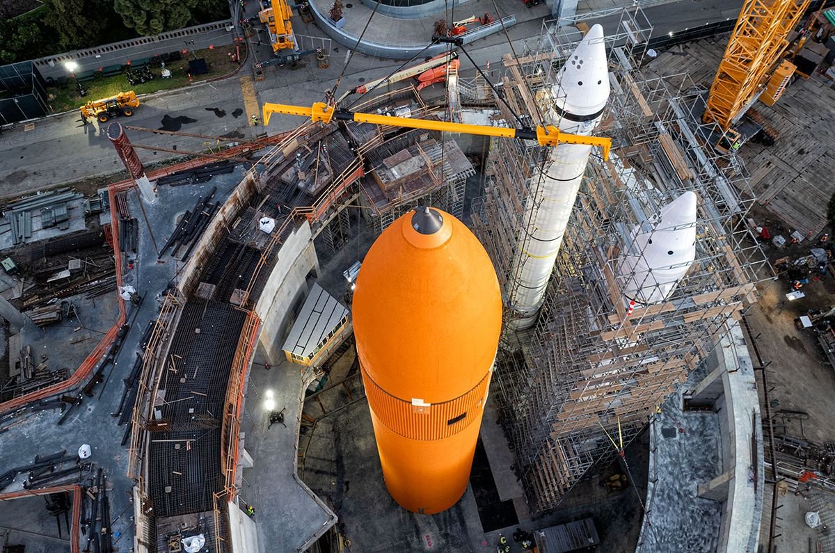 a large cylindrical orange fuel tank stands next to two large white rocket boosters which are surrounded by scaffolding