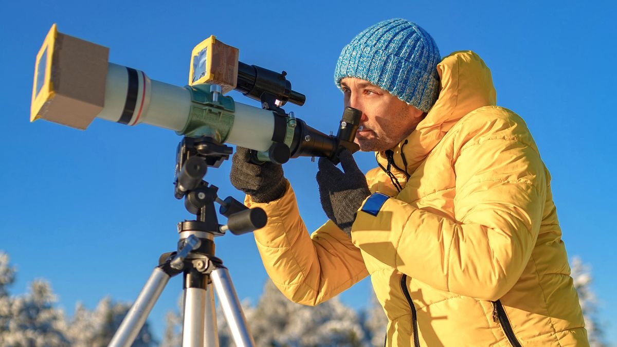 Male uses a telescope with solar filter to observe the sun in yellow jacket and with a blue sky behind