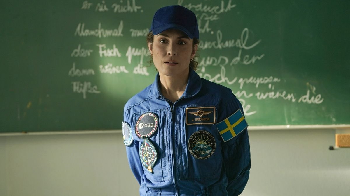 A woman wearing a blue astronaut jumpsuit decora ted with several different patchers including a Swedish flag She is also wearing a blue baseball cap She is standing in front of a black board with some chalk writing on