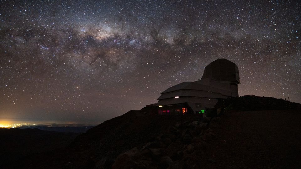 the silhouette of an observatory sits atop a dark hill beneath the milky way stretched across a starry sky