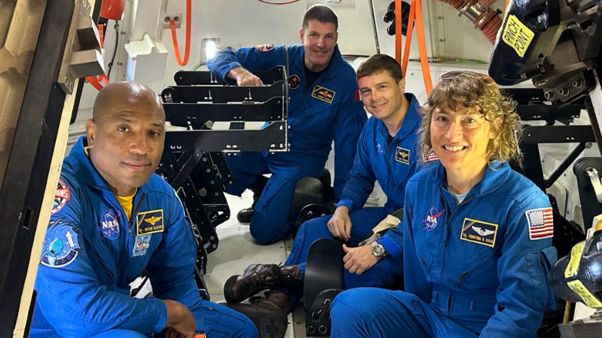 four astronauts in a small spacecraft squatting and smiling at the camera while wearing flight suits