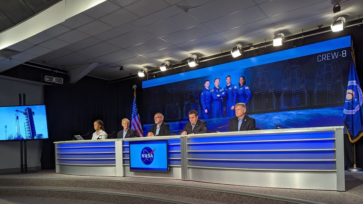 people in suits sit at a desk in front of a poster of four astronauts in flight suits and the text