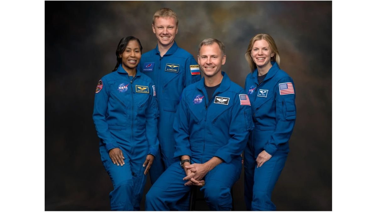 four smiling people in blue flight suits stand in front of a dark gray brown background
