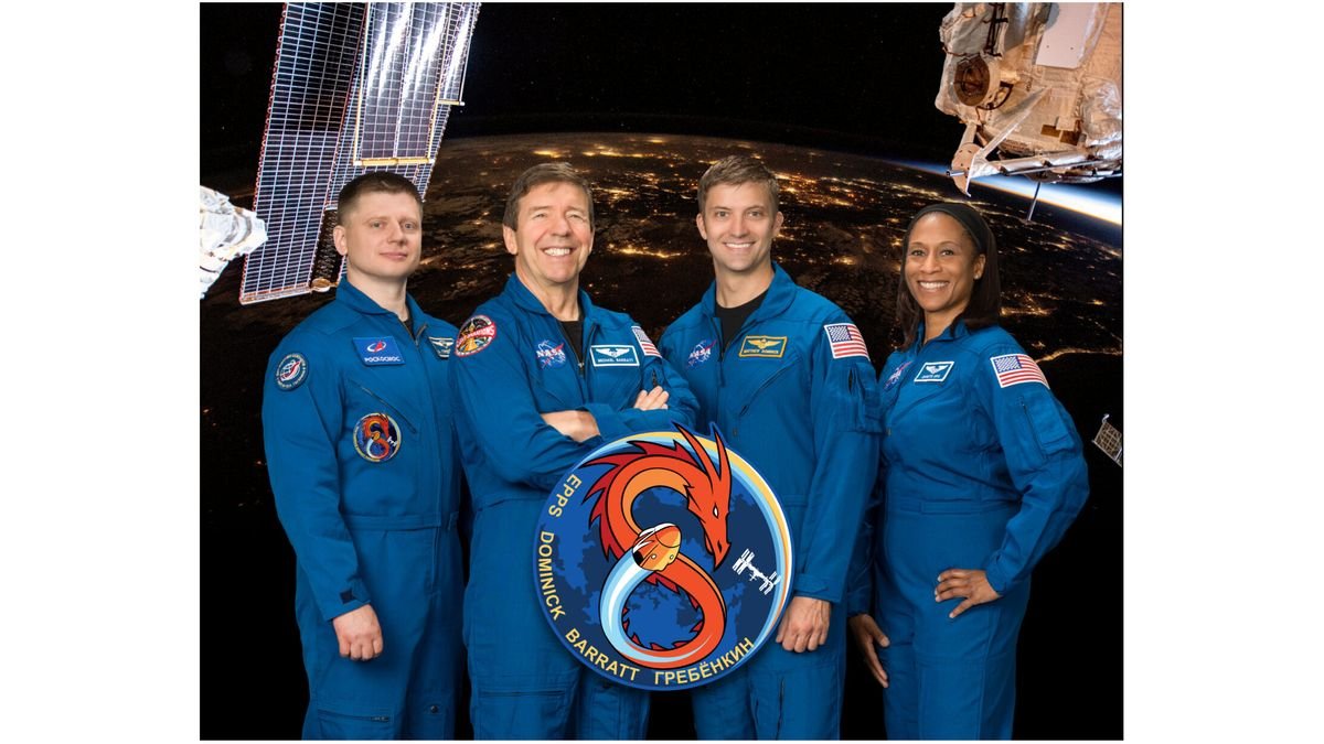 four smiling people three men and one woman in blue flight suits stand in front of an image of earth at night captured from low earth orbit