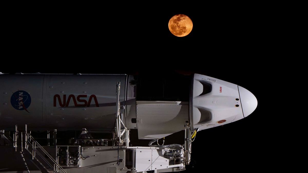 a white rocket and space capsule are transported to the launch pad horizontally at night with the yellow orange moon just above them in the dark sky