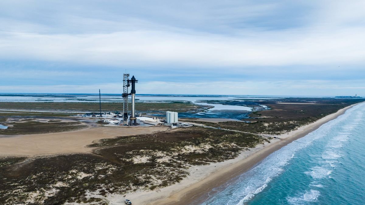 a huge silver and black rocket stands amid sand dunes on a seaside launch pad