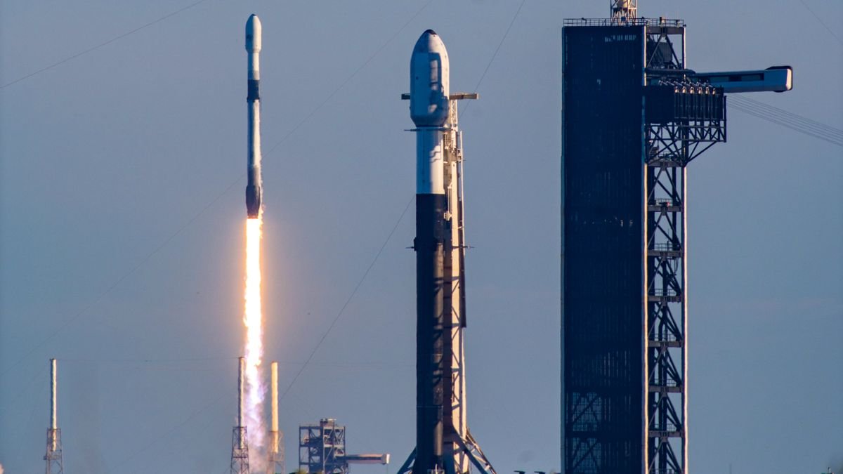 a white and black spacex falcon 9 rocket launches into a blue sky behind another falcon 9 which is poised on a launch pad ready for liftoff