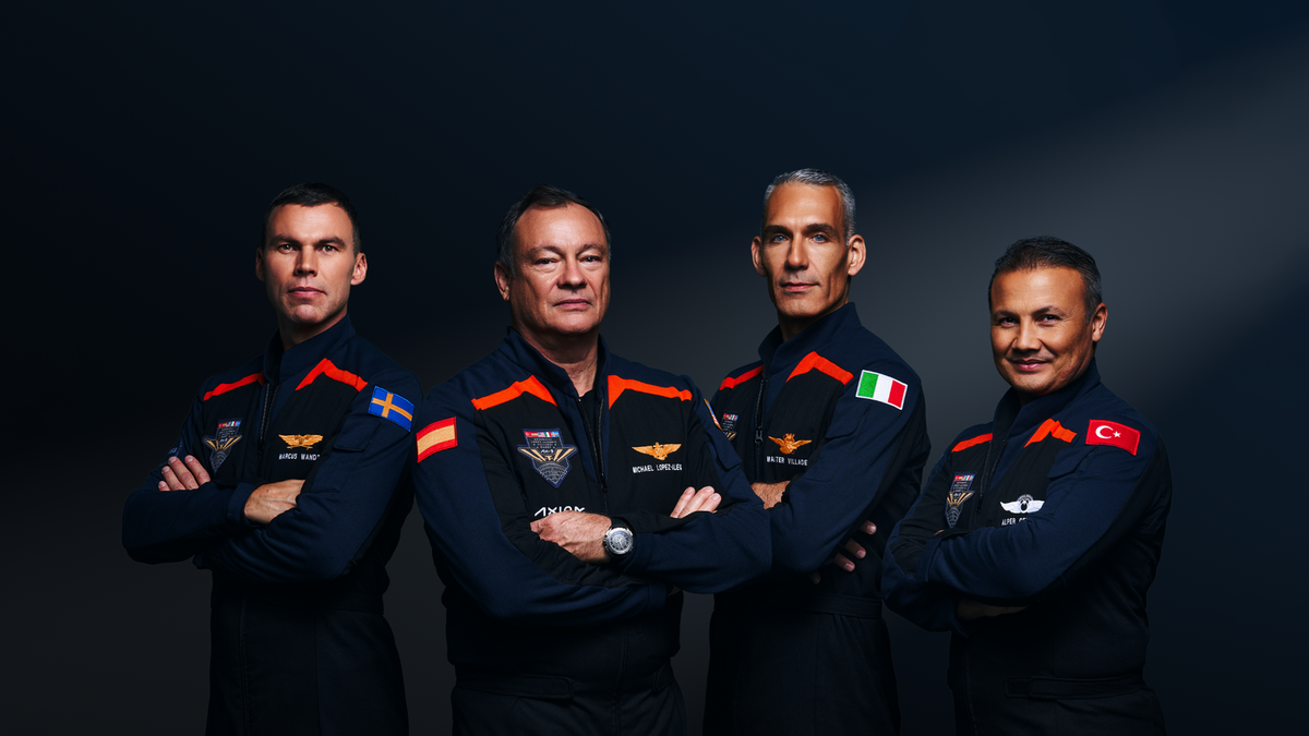 Four men stand with arms crossed wearing dark blue jumpsuits with red accented shoulder wings They look serious except for the smiling man on the right He is shorter than the rest