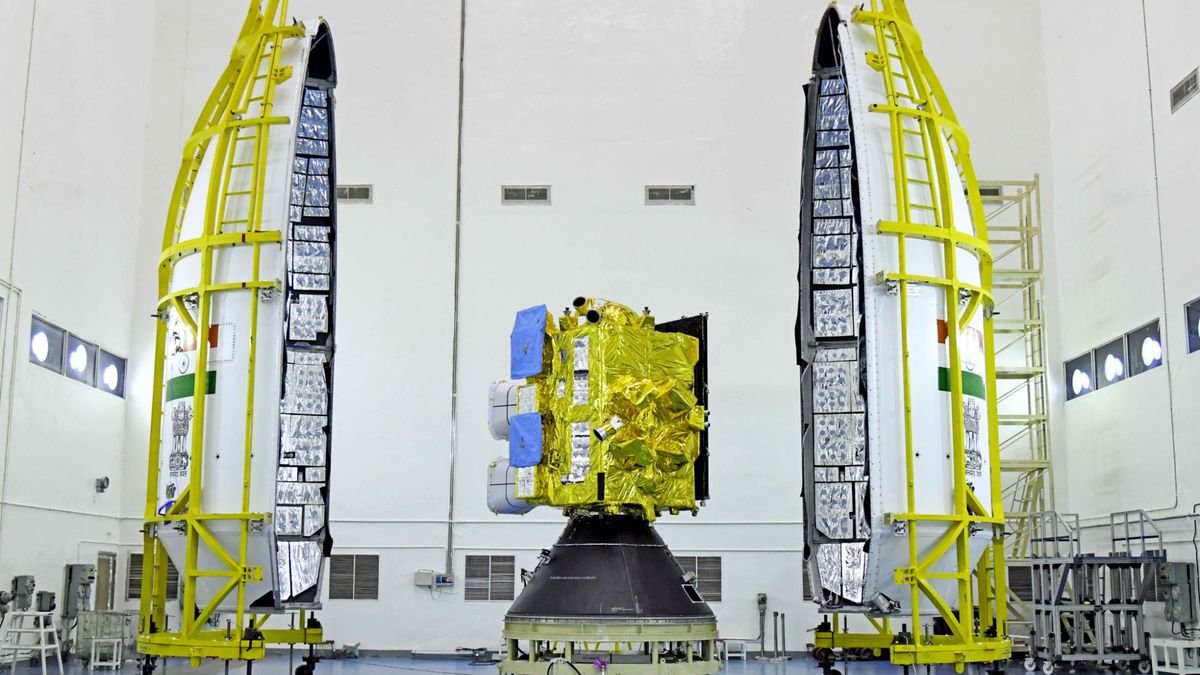 a cubical spacecraft wrapped in gold foil sits in a large white walled clean room between the two white halves of its bullet shaped payload fairing