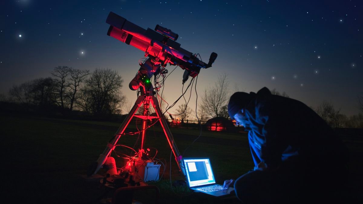 Somebody crouched down using their laptop in the night in front of their telescope