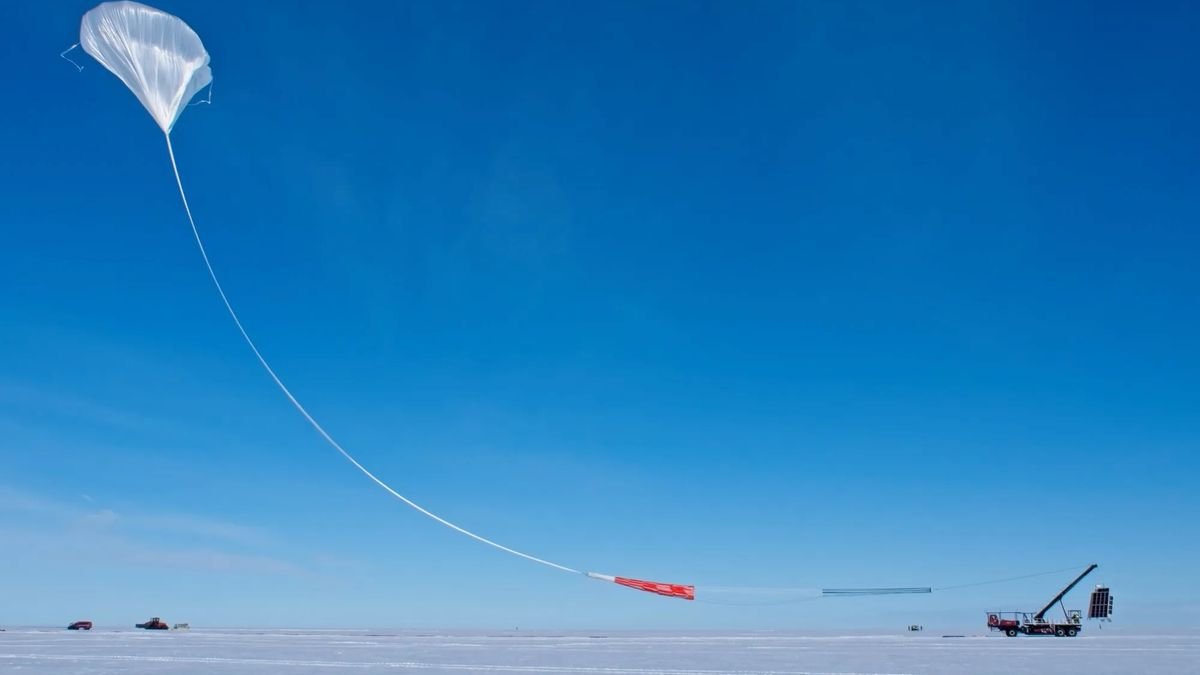a large white scientific balloon launches into a blue sky from a flat snow covered patch of ground