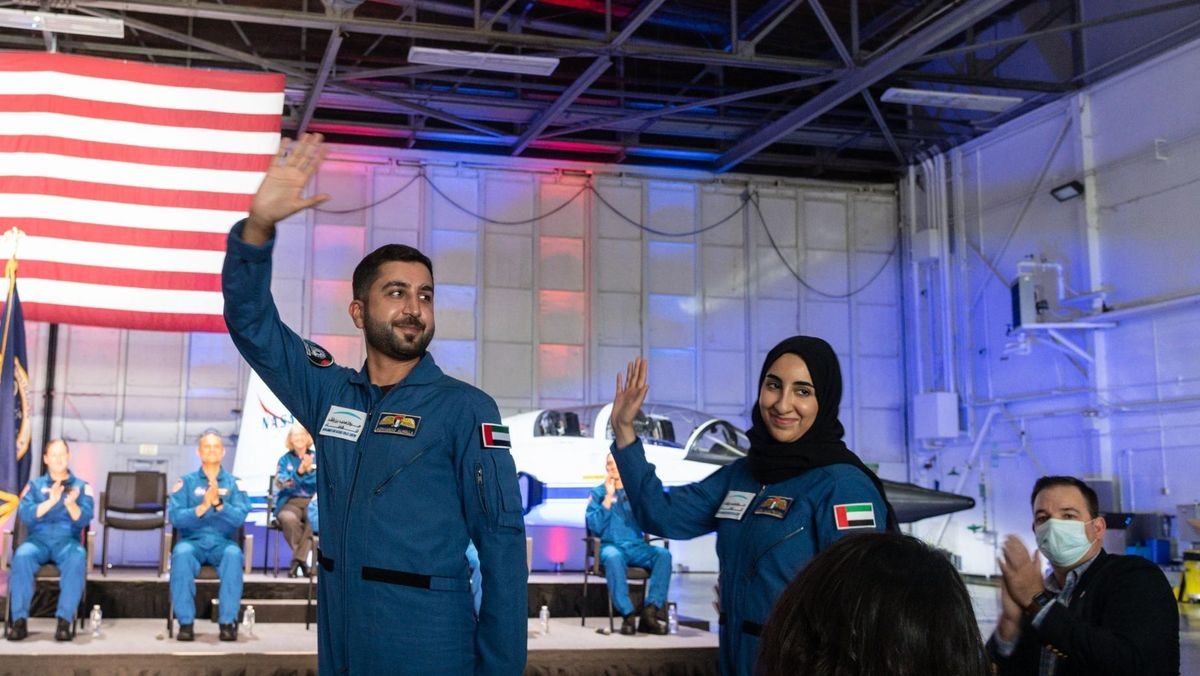 two astronaut candidates waving in flight suits in front of a us flag and a t 38 jet on the stage behind seated astronaut candidates applaud