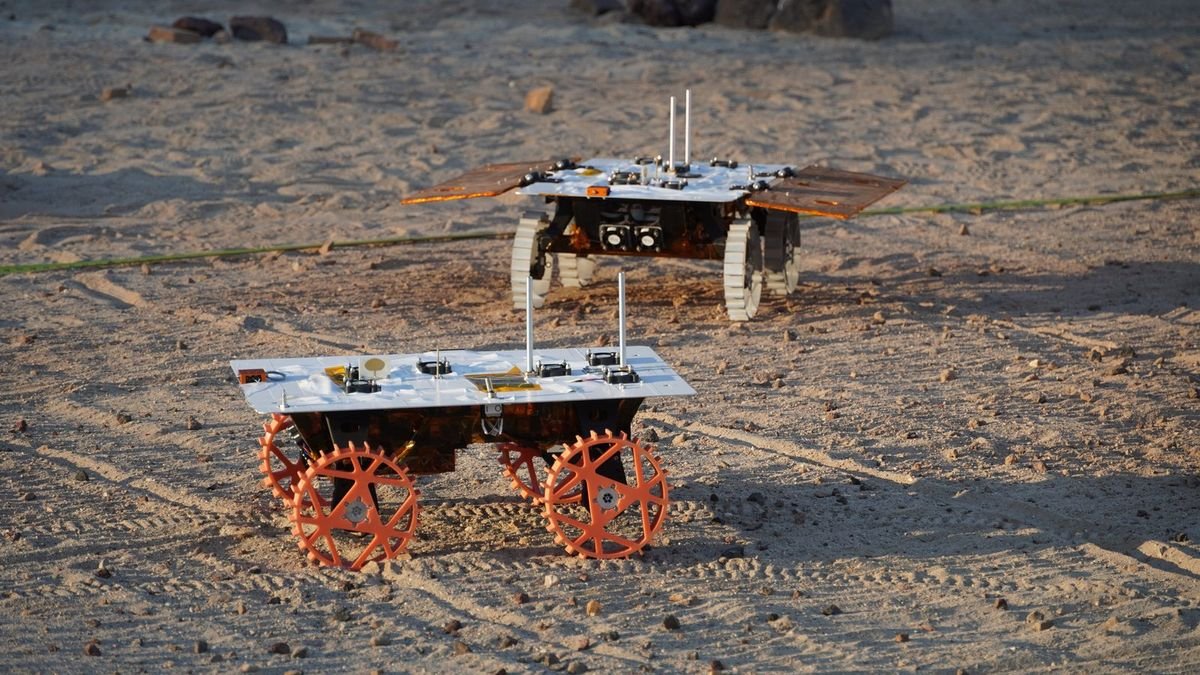 two small four wheeled robotic rovers drive in a sandy enclosure during a test