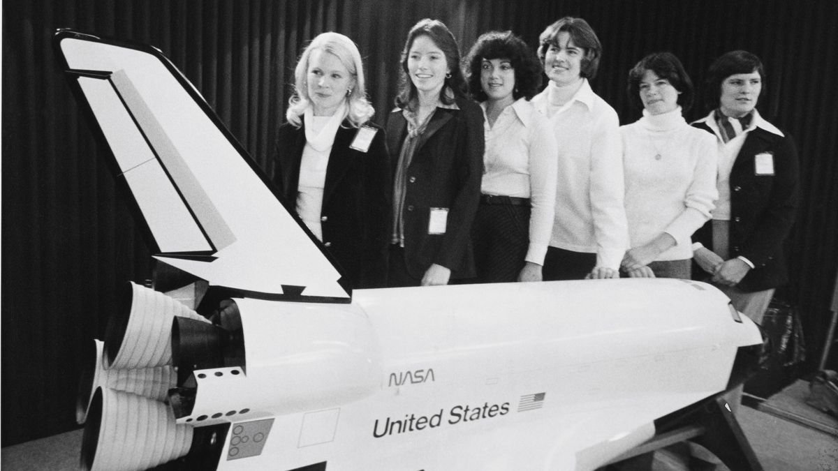 Six black and white women all white stand behind a giant model of the space shuttle