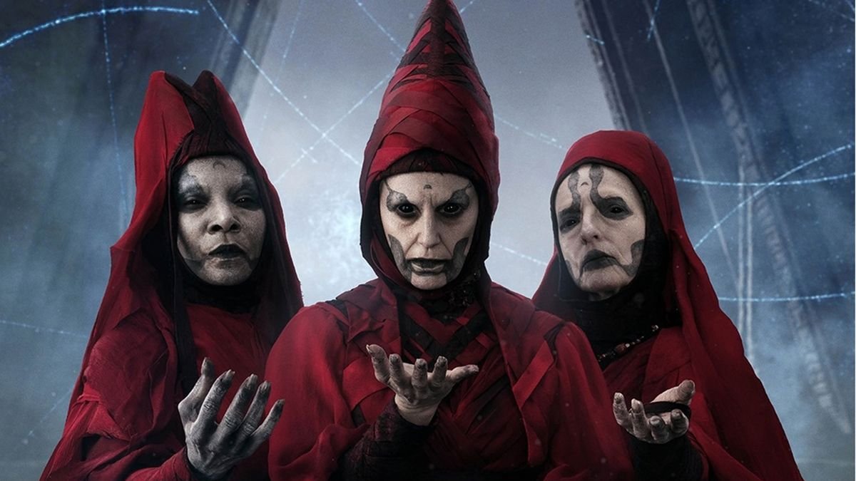 Three menacing women wearing dark red robes with hoods They have pale white faces with black markings They each have one hand raised palm up Image of some of the Witches of Dathomir from Star Wars