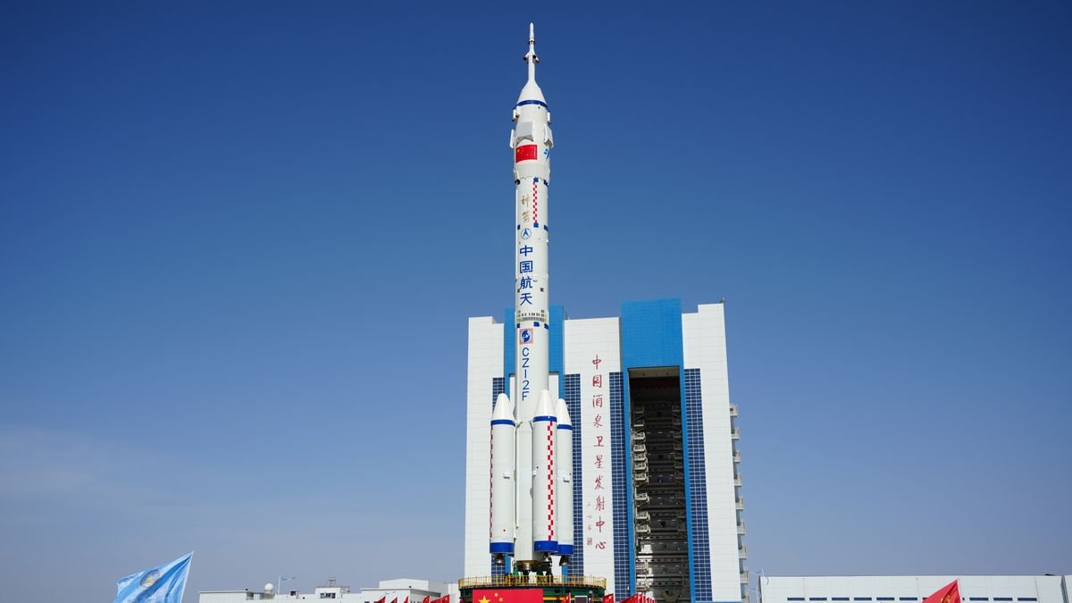 a tall white rocket with a red square near the top and chinese writing on the side stands outside a hanger building beneath a blue sky
