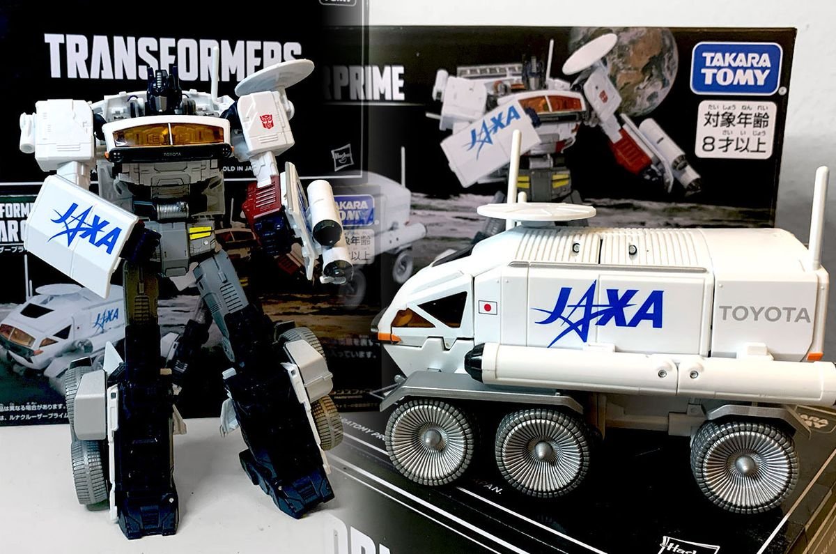closeup of a six wheeled white moon rover transformer toy with the words jaxa and toyota written on its side in front of the box it