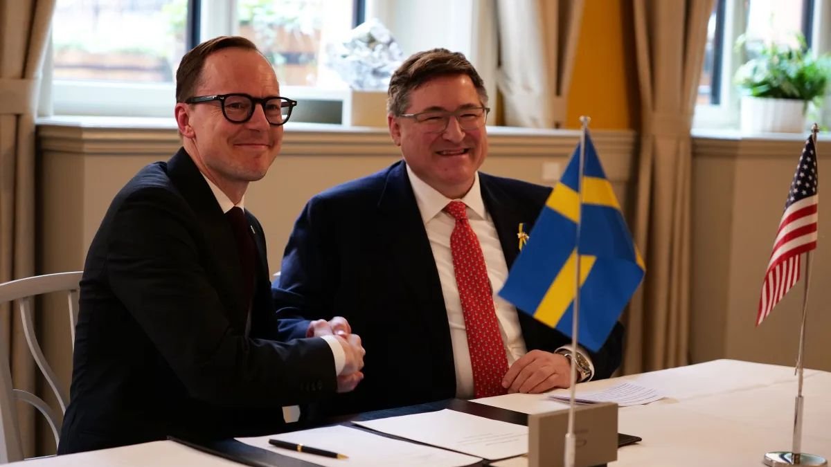 Two men in dark suits shake hands while sitting at a table with documents and two small flags Both men are wearing glasses