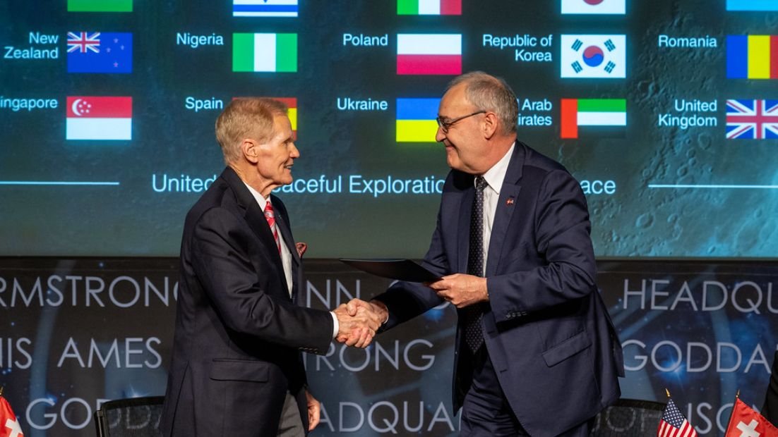two white men in dark suits shake hands in front of a backdrop of many flags