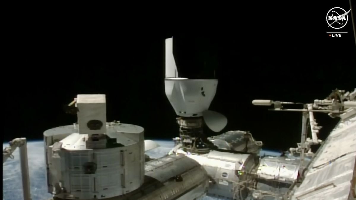 a spacecraft capsule is docked upside down to the upward docking port of a chrome space station module