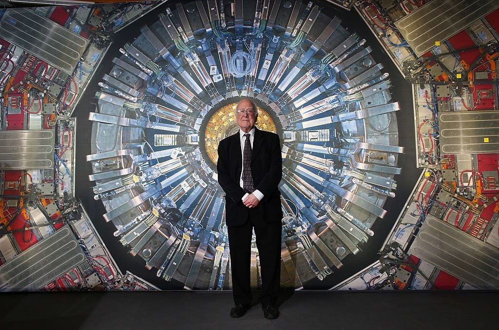 A man in a suit stands in front of an image of the Large Hadron Collider which has lots of mechanical parts centered around a yellow middle device