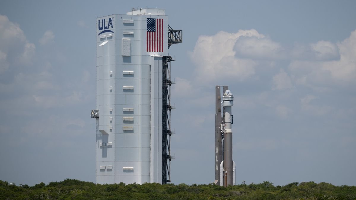 a brown and white rocket rolls vertically toward a large white building with cloudy blue skies in the background