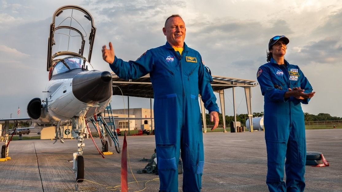 two astronauts in flight suits standing in front of a t 38 jet with cockpit open they are on a tarmac with a structure in behind and buildings far in the back