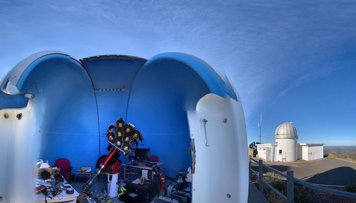 an open sided dome structure hovers above telescope equipment in the foreground behind to the right a domed silo with blue skies above