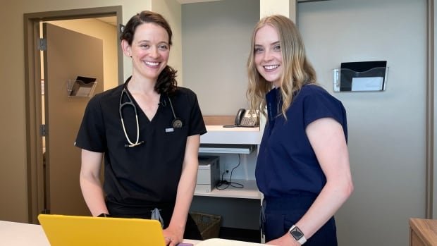 Julie Croteau and Jordan McPhee wear dark scrubs and stand in front of a yellow laptop in a clinic hallway
