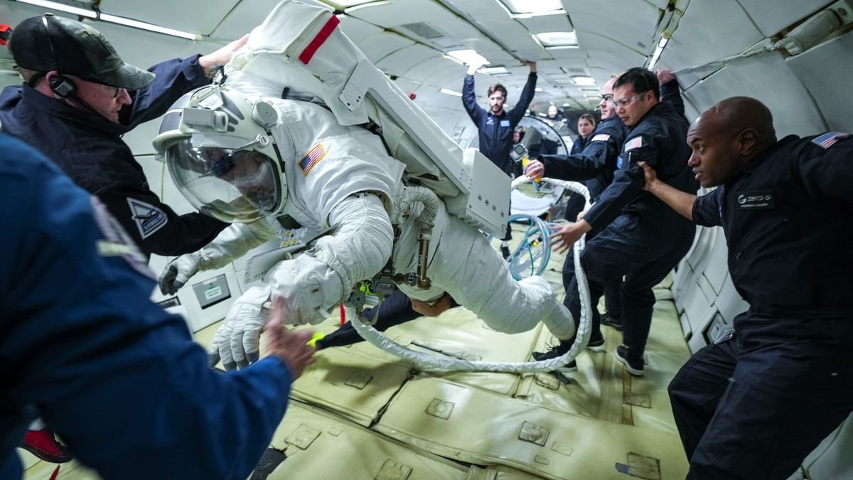 a person in a bulky spacesuit floats weightless inside the fuselage of an aircraft surrounded by people in blue flight suits and safety goggles