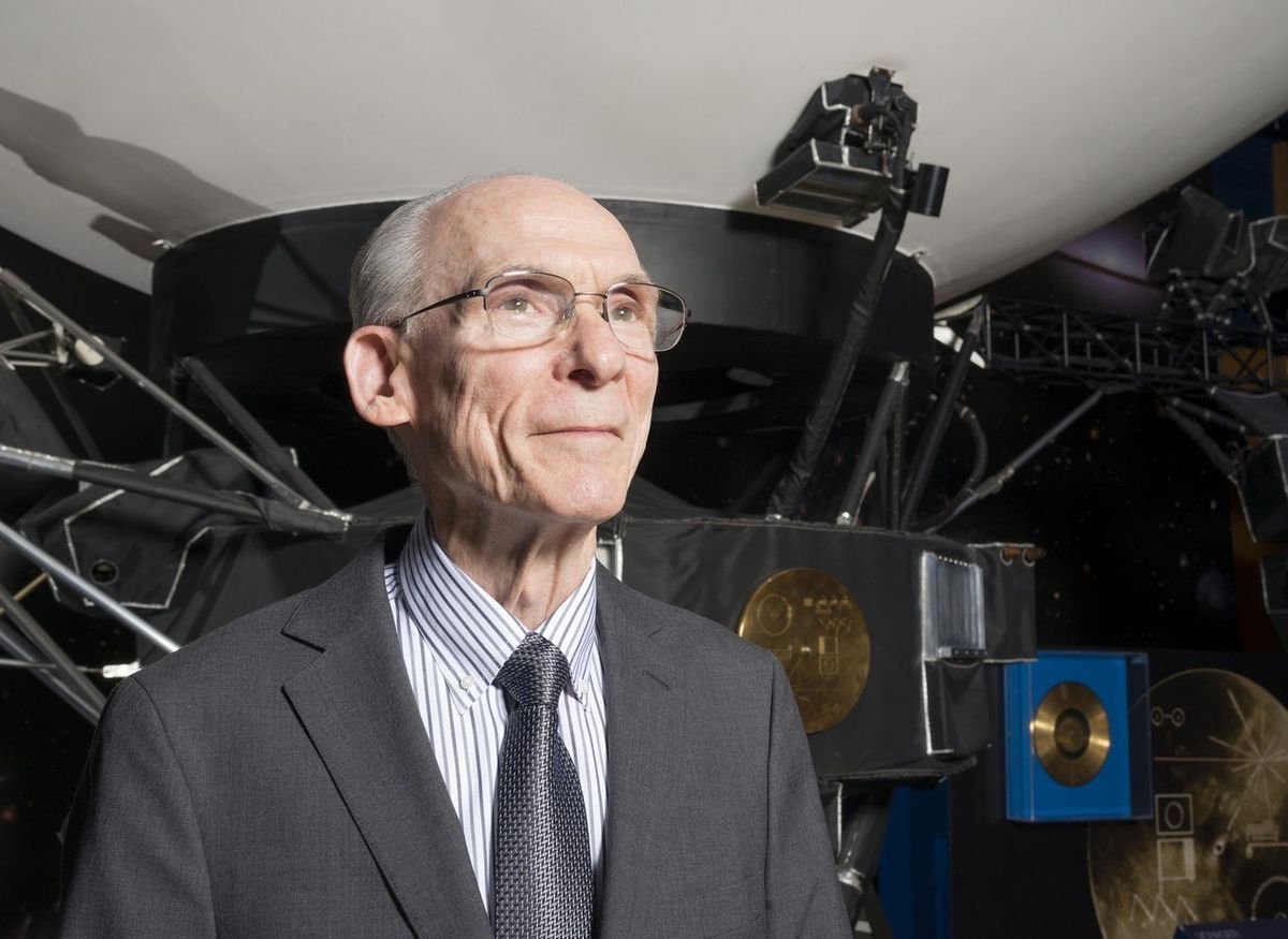 a smiling older man in a suit stands in front of a model of a large spacecraft