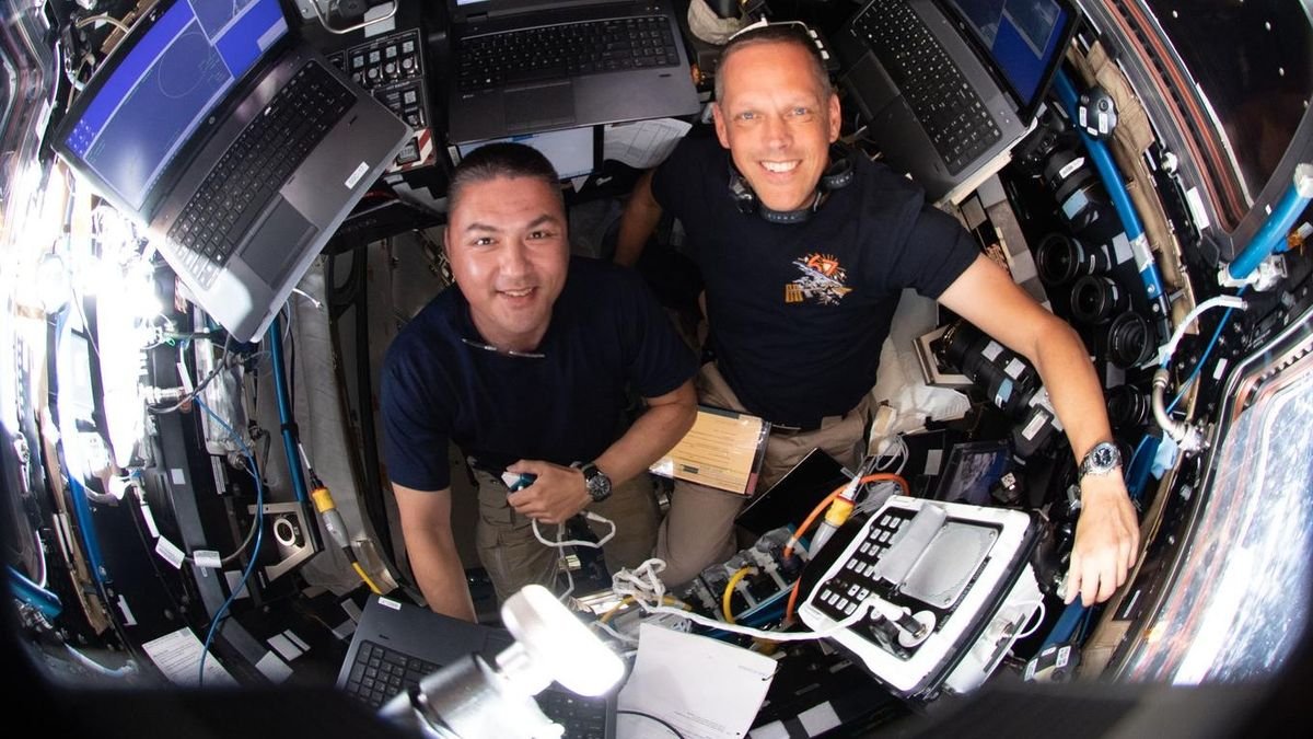 an overhead shot of two astronauts smiling in a small windowed room of the international space station surrounded by computers and a window