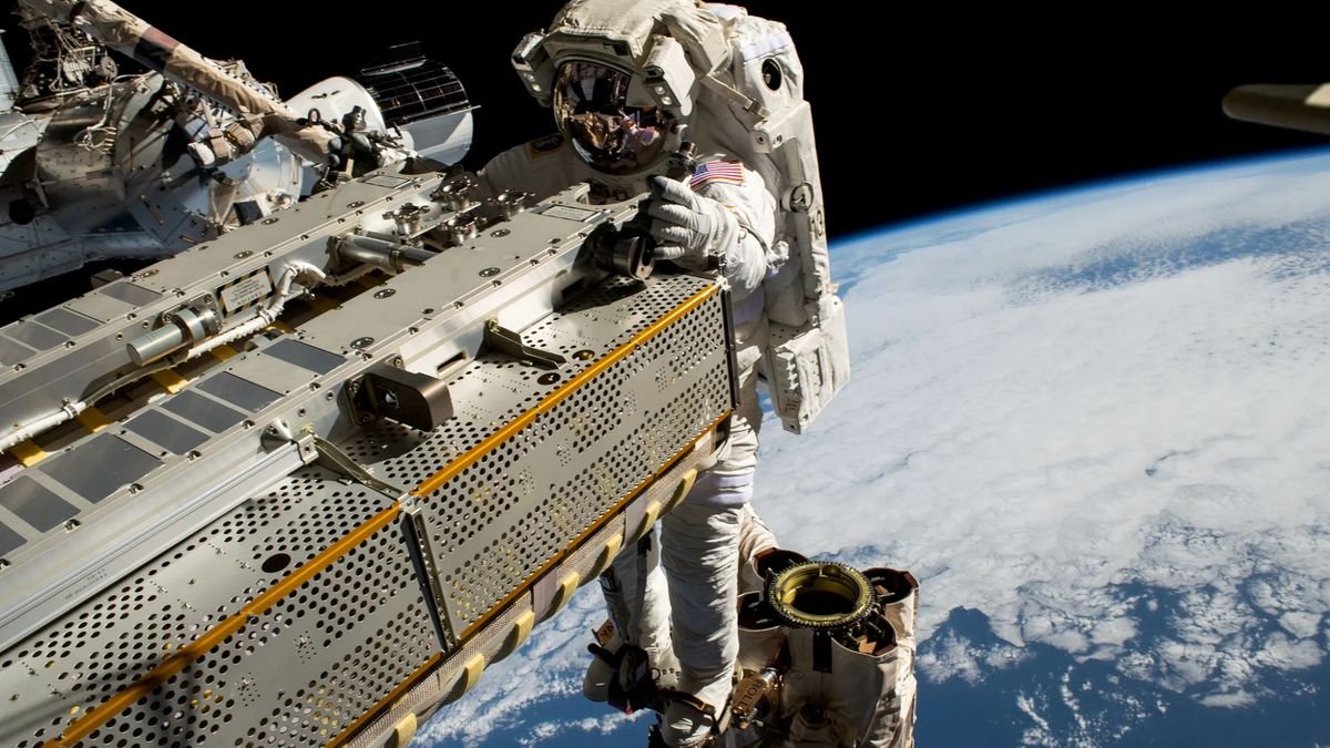 an astronaut in a white spacesuit grabs a piece of the international space station during a spacewalk with earth in the background