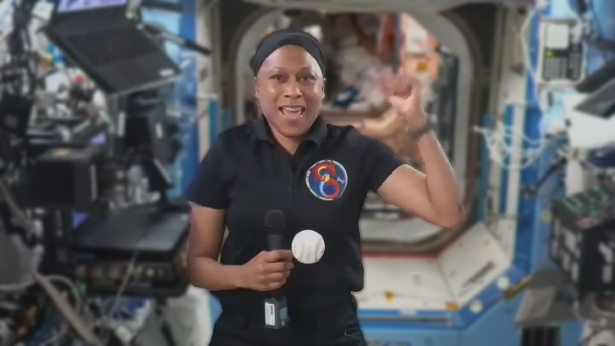 an astronaut waving their hand with a baseball in front while floating on the international space station computers are in the background as well as an open door