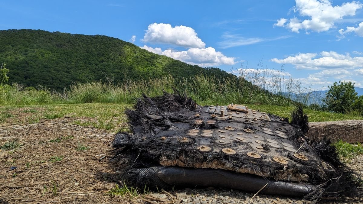 a large black piece of fiberglass covered in metal bolts and plates lies on the ground beside a trail leading into a forest mountains can be seen rolling in the distance