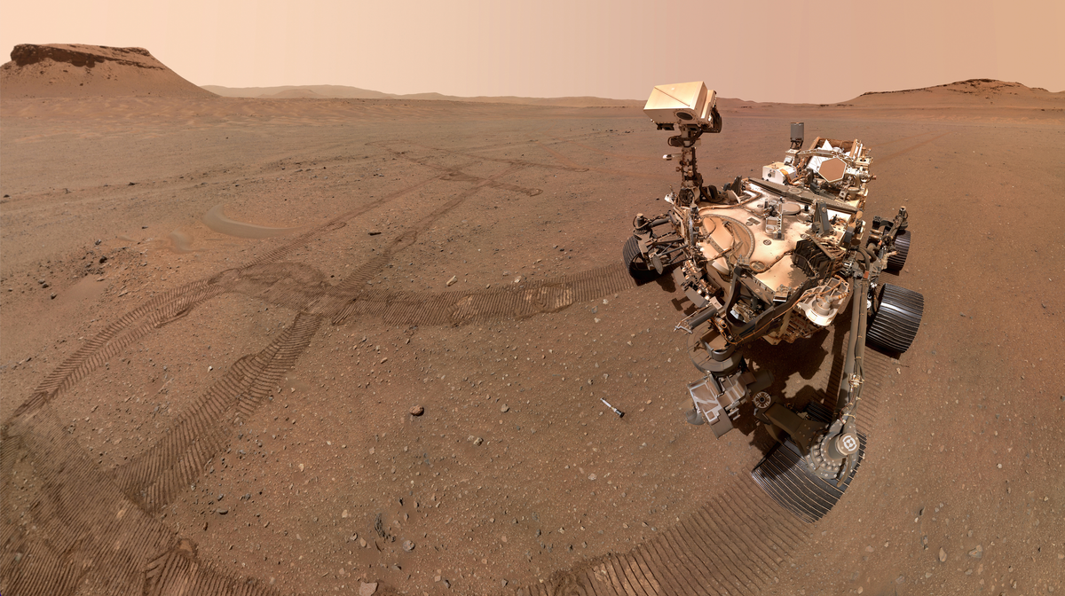 a four wheeled robot with a head like box atop a neck like appendage looks down at a silver tube lying on the dusty reddish orange surface of Mars
